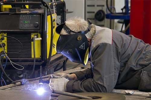 A welder in a helmet laying a bead on steel tubing at a workshop bench